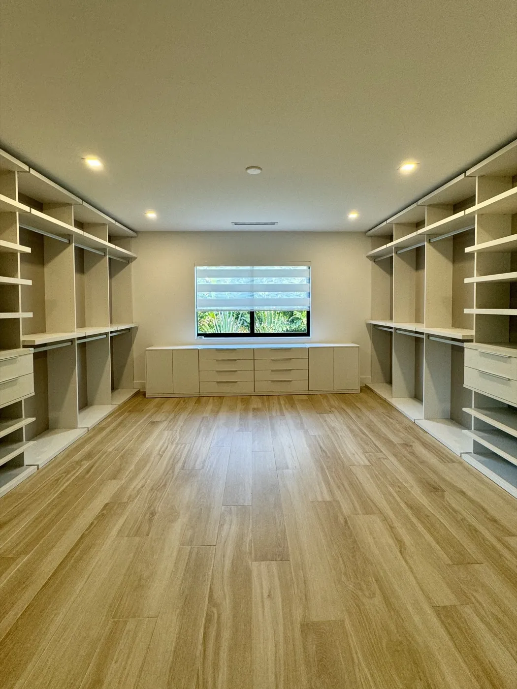 Walk-in closet with built-in shelving and hanging rods on both sides, drawers under a window and wood-look porcelain plank flooring