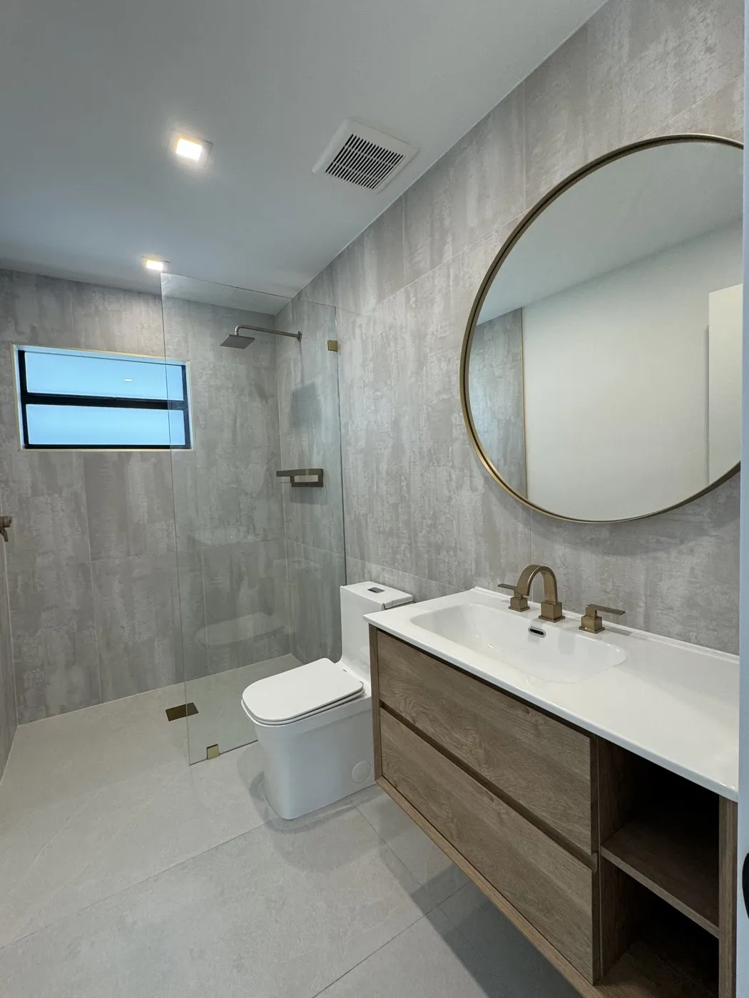 Bano completo nuevo en un antiguo cuarto de lavado con ducha walk-in, panel de vidrio sin marco, tocador flotante de madera y espejo redondo dorado