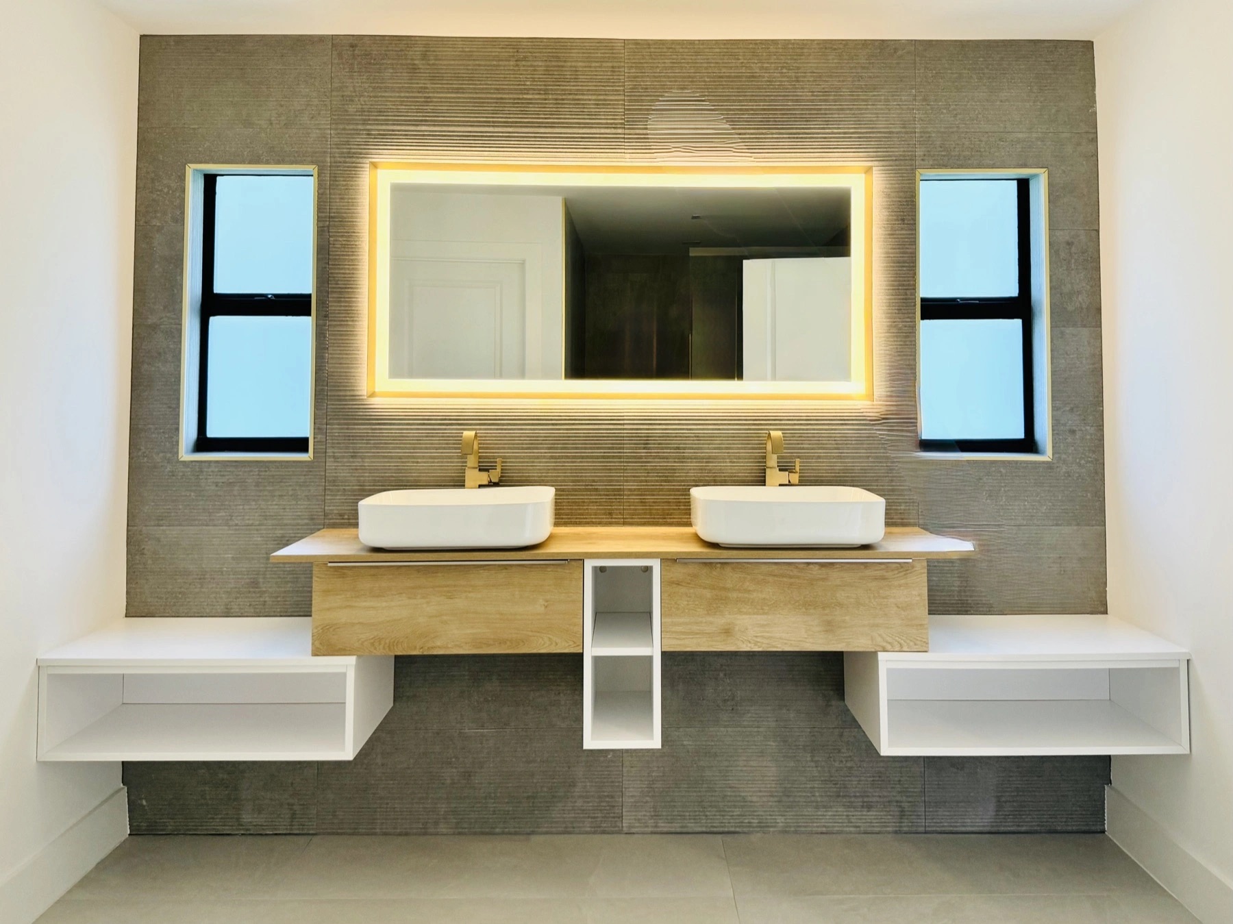 Bano principal remodelado en Fort Myers, FL con tocador doble flotante, lavamanos de sobreponer blancos, griferia dorada cepillada, espejo LED retroiluminado y pared de azulejo texturizado